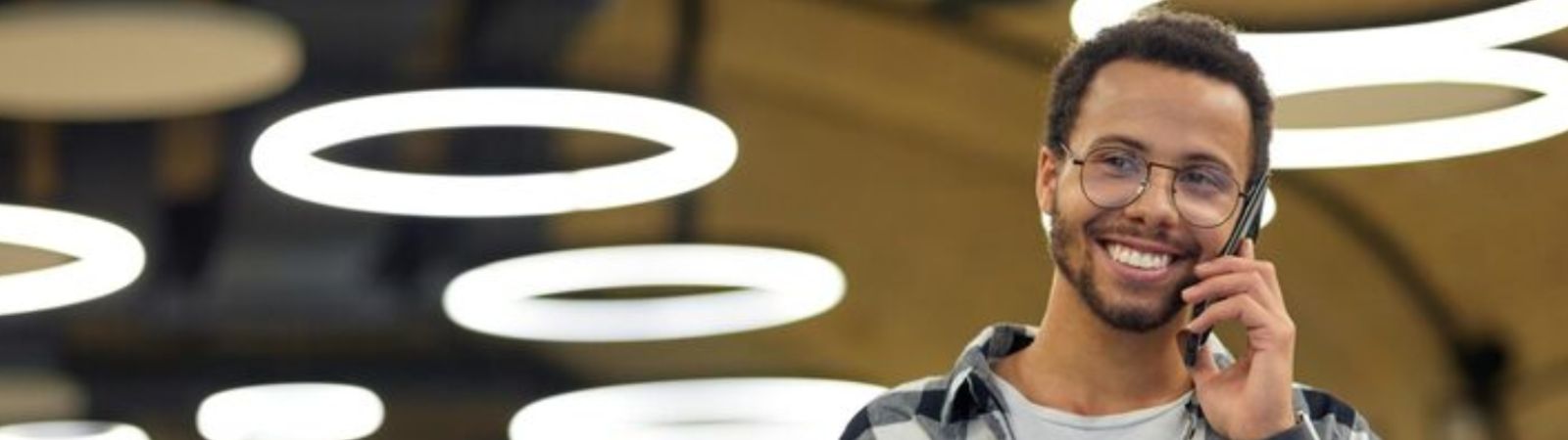 Homem negro sorrindo com óculos e fone de ouvido em ambiente interno com luzes circulares brancas no fundo falando ao celular.
