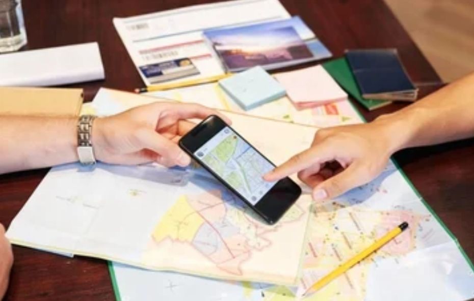 A imagem exibe uma visão aproximada de uma mesa de trabalho onde duas pessoas planejam uma rota. Sobre a mesa, há um mapa impresso detalhado, blocos de notas coloridos, canetas e materiais impressos que sugerem guias de viagem. Uma das mãos segura um smartphone que exibe um mapa digital, enquanto a outra aponta para um local específico na tela, simbolizando a integração entre o planejamento analógico e as ferramentas digitais de geolocalização.