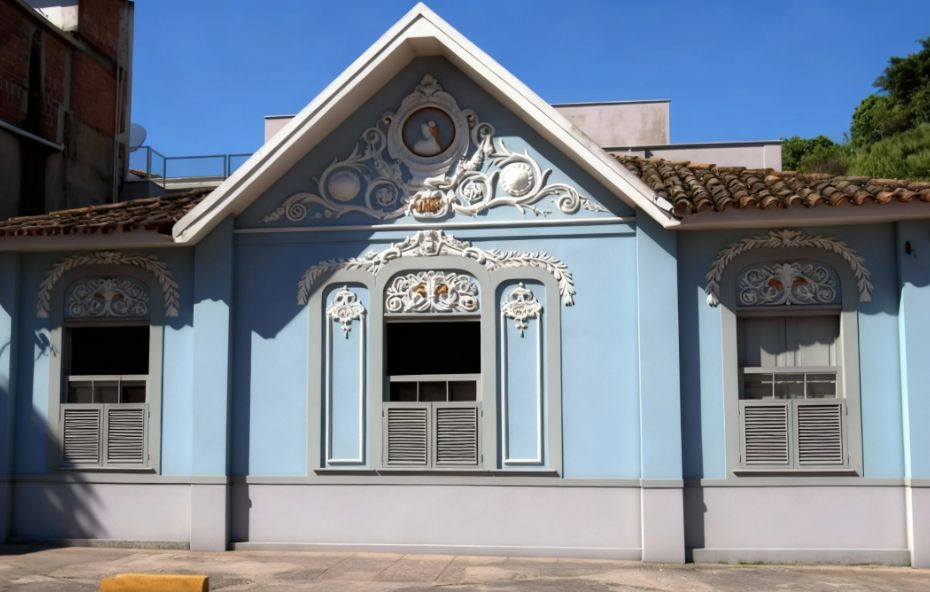 Fotografia da fachada de um casarão histórico de cor azul clara com detalhes arquitetônicos em branco e telhado de barro marrom. A imagem mostra um frontão central elevado de formato triangular que abriga um medalhão oval com a imagem da Princesa Isabel e a inscrição do ano de mil oitocentos e sessenta e quatro. Abaixo do frontão há uma janela central tripla e janelas laterais adornadas com guirlandas e ornamentos clássicos esculpidos em relevo.
