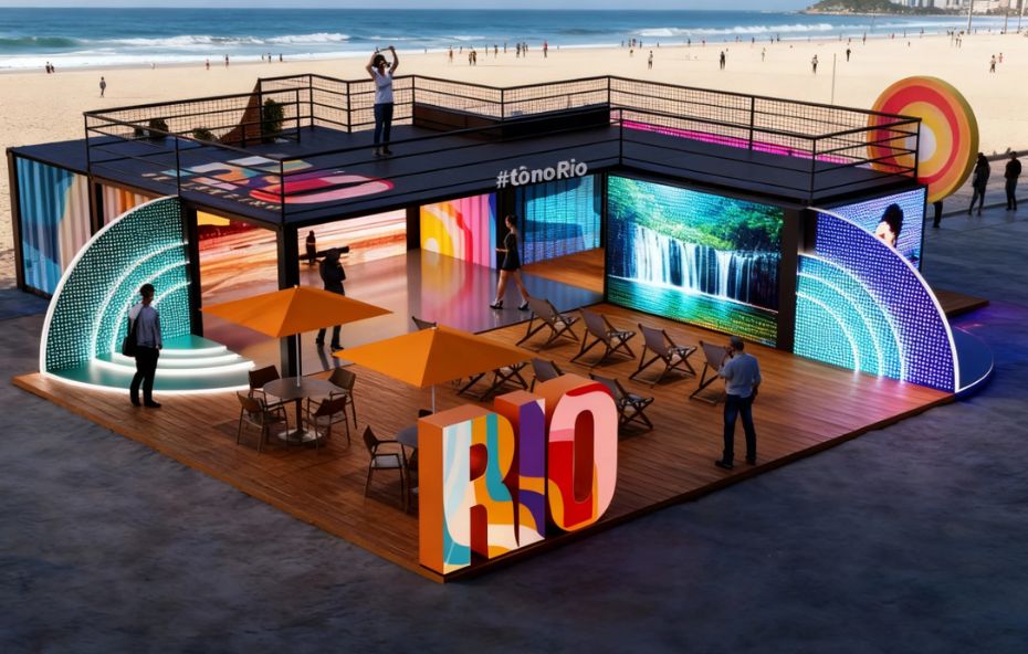 Fotografia aérea de um estande moderno feito de contêineres pretos na areia da Praia de Copacabana. A estrutura possui dois andares com terraço superior e escada lateral iluminada em LED verde. No deck de madeira frontal existem ombrelones laranjas e cadeiras de praia. Um grande letreiro colorido com a palavra RIO se destaca no centro. Telões de LED exibem imagens de uma cachoeira e artes abstratas coloridas. Pessoas caminham pelo espaço sob a luz do sol.