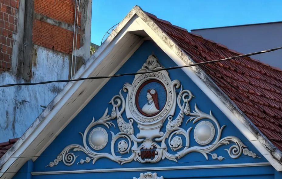 Fotografia em close do frontão triangular de uma edificação histórica pintada em azul vibrante com ornamentos brancos em relevo. No centro do triângulo há um medalhão oval com a pintura de perfil da Princesa Isabel ladeado por guirlandas de flores esculpidas. Logo abaixo do frontão destaca-se o ano de mil oitocentos e sessenta e quatro pintado em branco sobre o fundo azul.