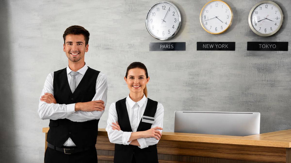 Um recepcionista homem e uma recepcionista mulher sorriem com braços cruzados atrás de um balcão de madeira com computadores. Na parede cinza ao fundo, há três relógios analógicos indicando os fusos horários de Paris, Nova York e Tóquio.