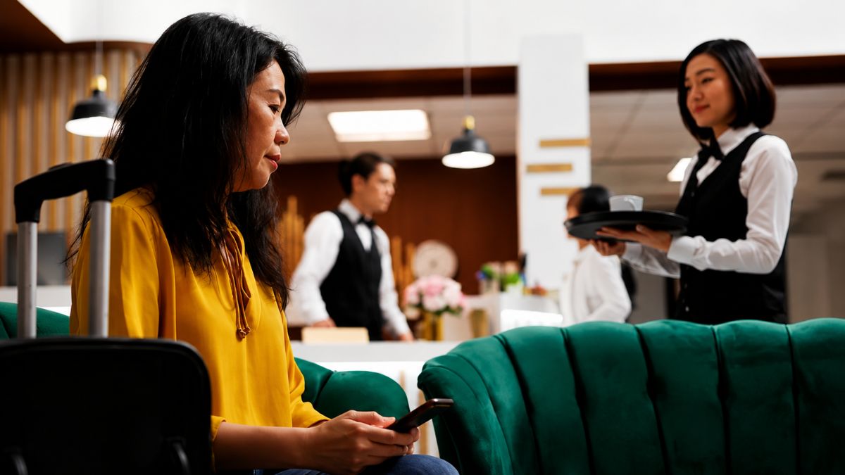 Hóspede sentada na recepção de um hotel utilizando o celular enquanto uma atendente transporta uma bandeja em um ambiente moderno e acolhedor.