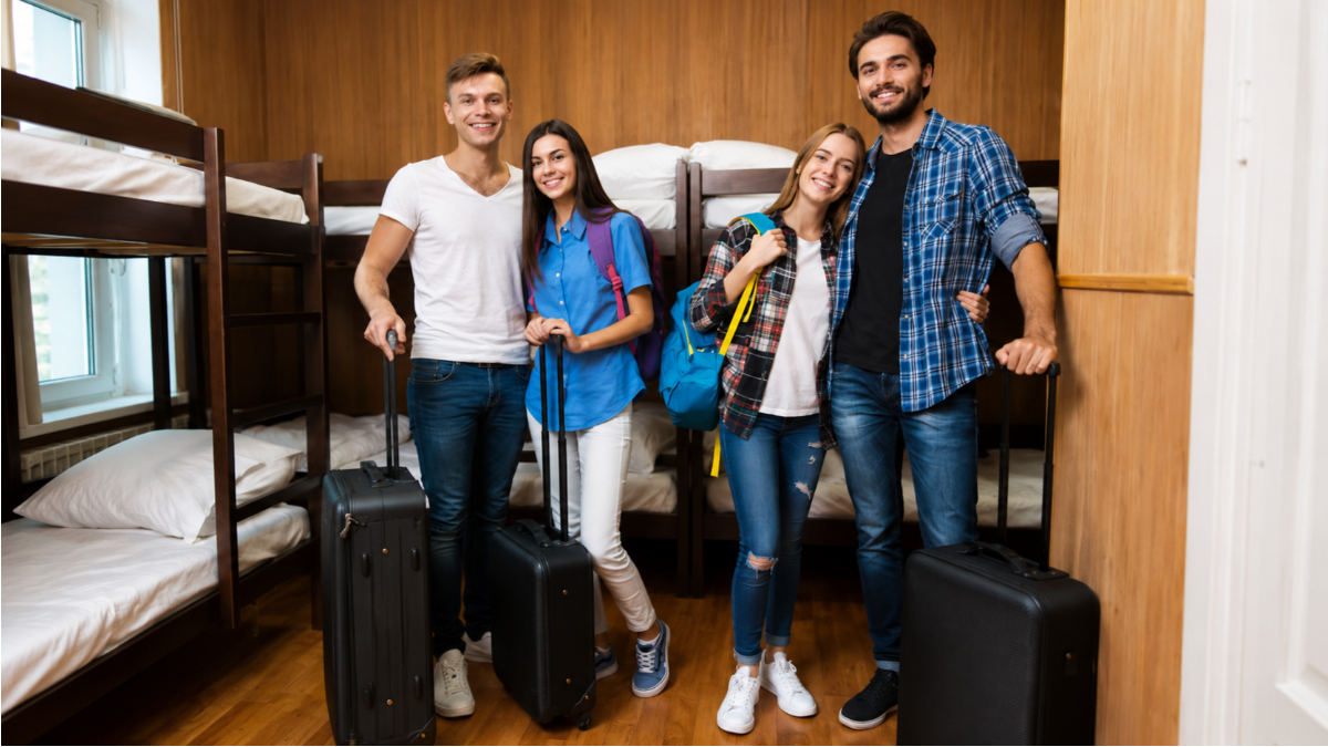 Quatro jovens viajantes, sendo dois homens e duas mulheres vestindo roupas casuais, estão em pé sorrindo e segurando malas de rodinha pretas dentro de um quarto coletivo equipado com beliches de madeira e lençóis brancos arrumados.
