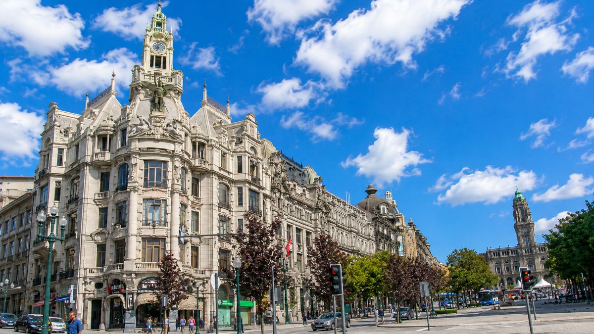 Edifício histórico com arquitetura detalhada e torre de relógio na Avenida dos Aliados com carros circulando e a Câmara Municipal do Porto ao fundo sob céu azul com nuvens na Baixa do Porto em Portugal.