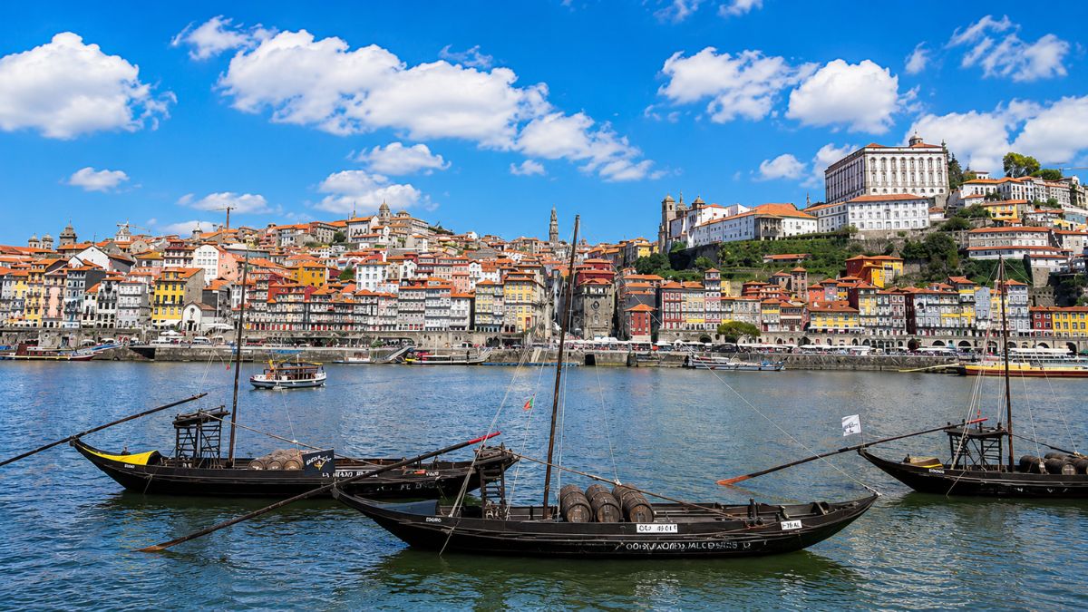 Barcos rabelos tradicionais ancorados nas águas calmas do Rio Douro com a colina histórica do bairro da Ribeira e suas casas coloridas ao fundo sob um céu azul na cidade do Porto em Portugal.