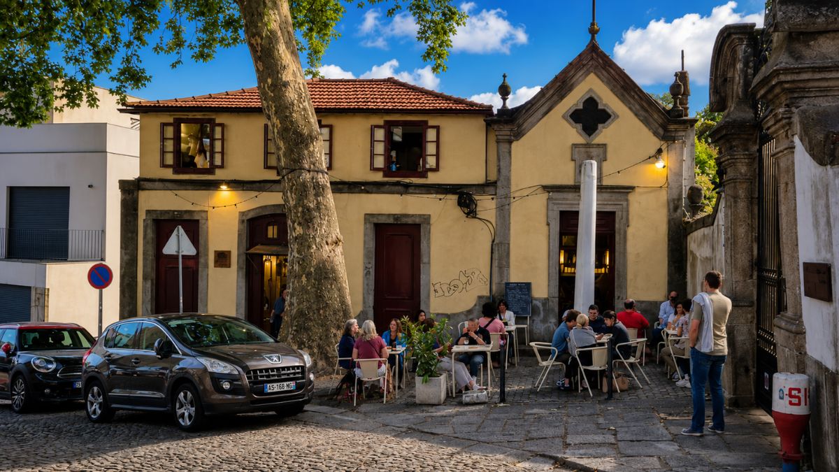 Pessoas sentadas em mesas de uma esplanada ao ar livre em frente a um edifício histórico amarelo com arquitetura tradicional e uma grande árvore no bairro de Cedofeita no Porto em Portugal sob céu azul.