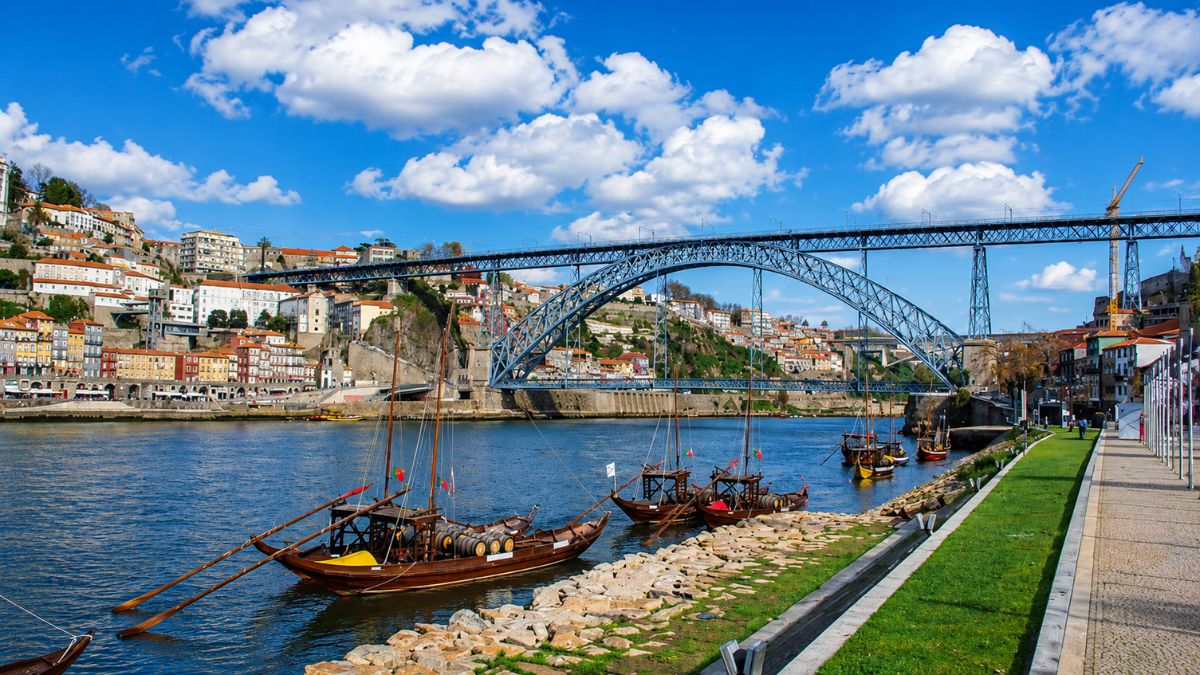 Vista da Ponte Luís I sobre o Rio Douro com barcos rabelos tradicionais ancorados na margem de Vila Nova de Gaia e o casario colorido do Porto ao fundo sob céu azul com nuvens.