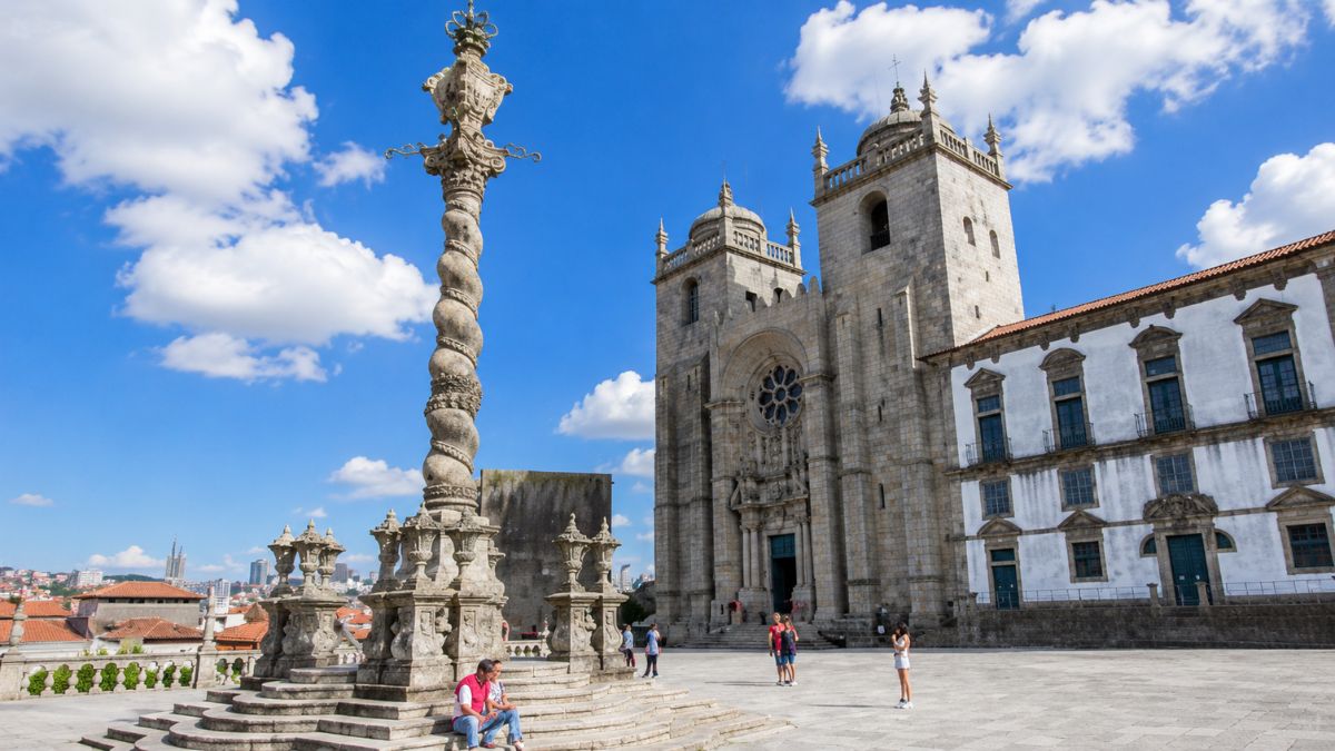 Vista da fachada de pedra da Sé do Porto com suas duas torres medievais ao lado do imponente pelourinho de estilo manuelino esculpido em espiral no centro da praça sob céu azul.
