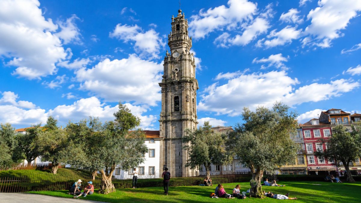Vista da imponente Torre dos Clérigos em estilo barroco erguendo-se atrás do Jardim das Oliveiras com pessoas sentadas no gramado sob céu azul com nuvens.