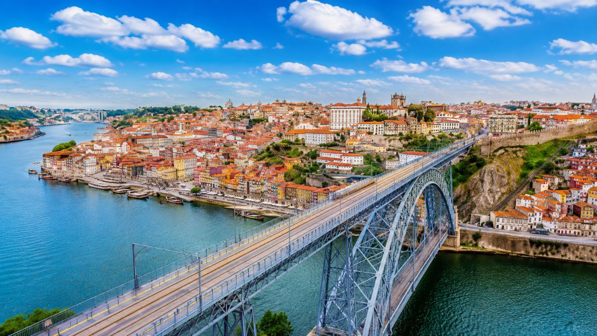 Ponte de ferro de dois tabuleiros sobre o Rio Douro com vista para as casas coloridas do centro histórico da cidade do Porto sob céu azul com nuvens.