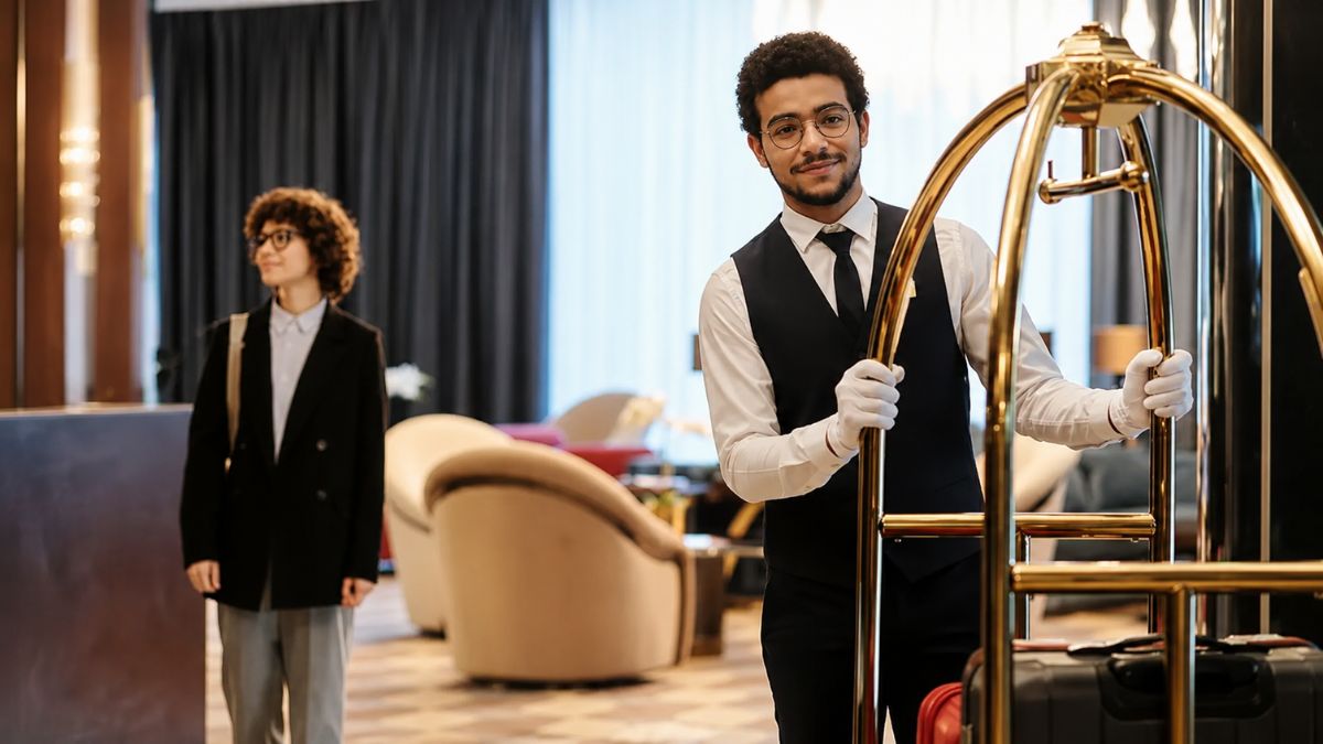Mensageiro de uniforme social escuro com luvas brancas segura um carrinho de bagagem dourado no lobby luxuoso de um estabelecimento hoteleiro.