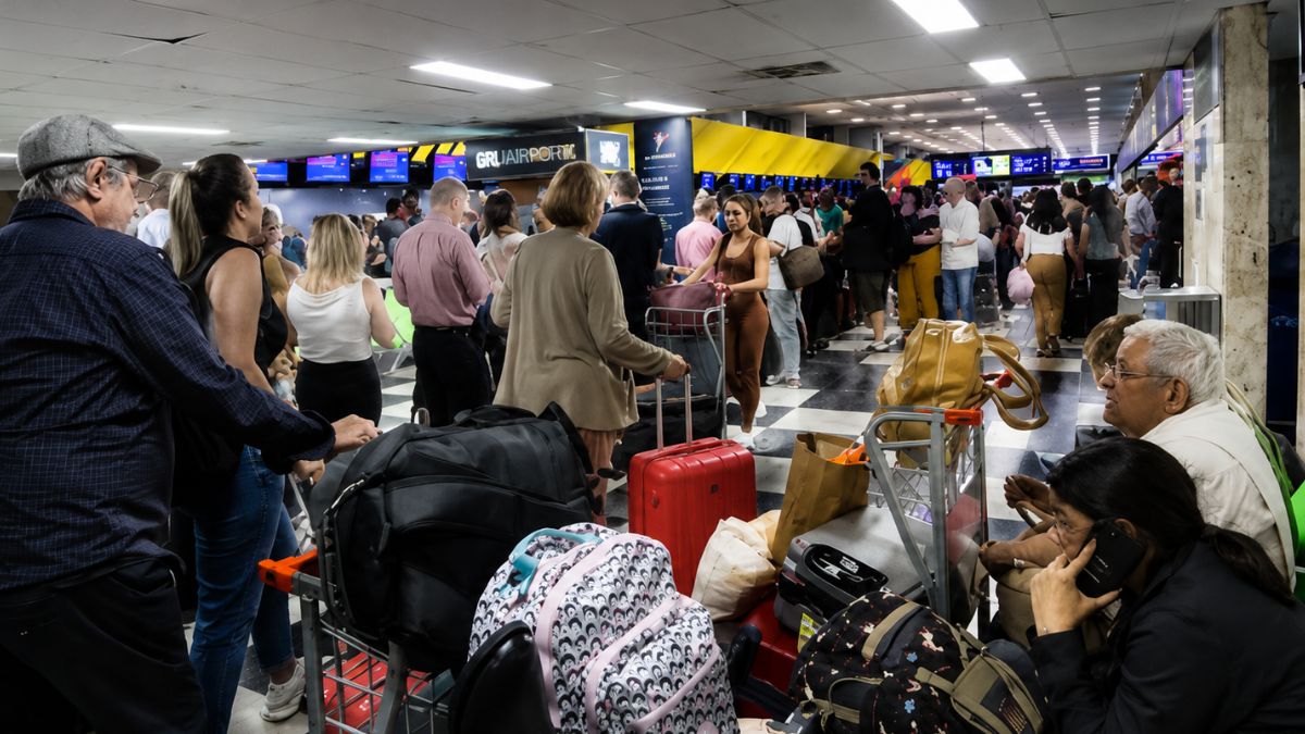 Fotografia colorida mostrando o saguão interno do Aeroporto de Guarulhos cheio de pessoas com malas e carrinhos de bagagem em filas diante dos balcões de atendimento das companhias aéreas.