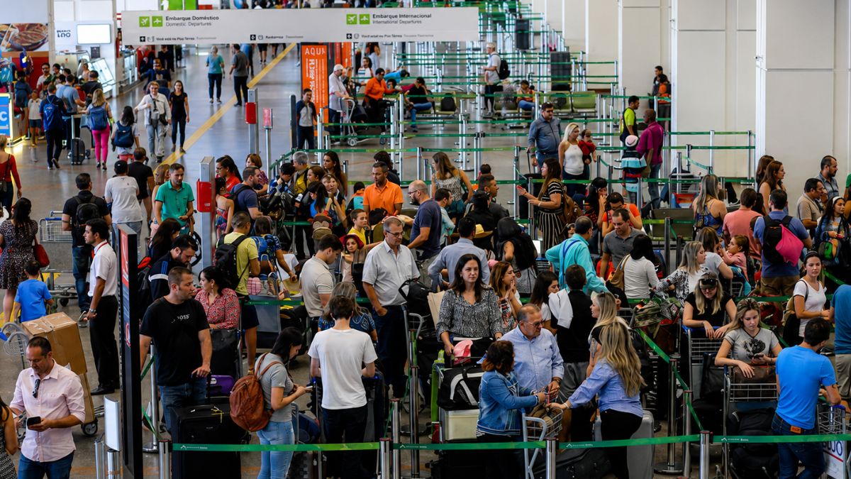 Fotografia em ângulo superior mostrando o saguão movimentado de um aeroporto com dezenas de pessoas carregando malas e mochilas organizadas em filas delimitadas por fitas organizadoras verdes diante dos balcões de atendimento ao fundo e placas de sinalização suspensas indicando embarque doméstico e internacional.