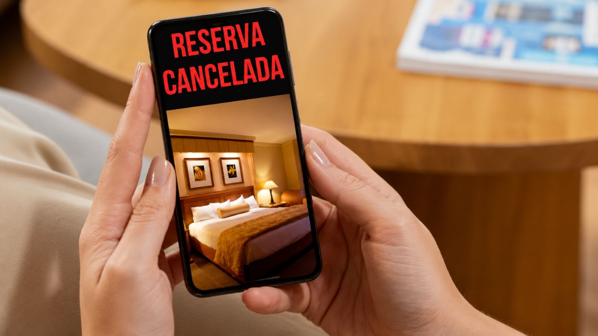 Mãos de uma pessoa segurando um smartphone na vertical que mostra a foto de um quarto de hotel luxuoso com iluminação aconchegante e um texto em destaque vermelho escrito reserva cancelada no topo do visor.