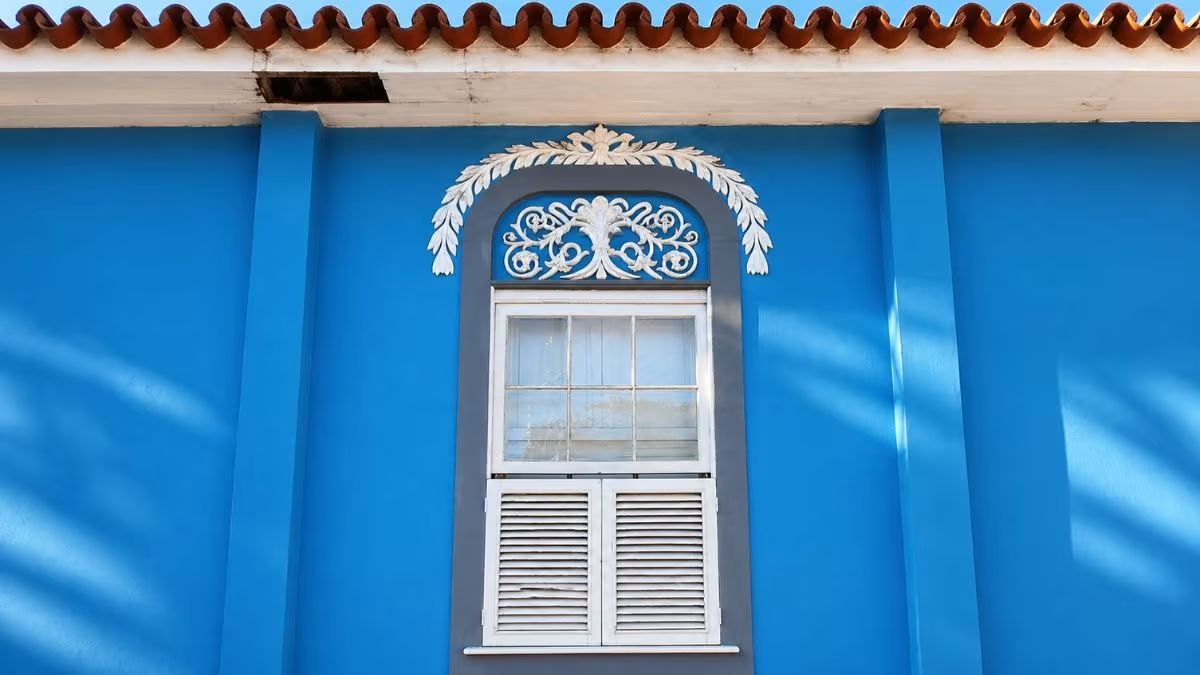 Janela retangular com venezianas brancas e moldura cinza emoldurada por guirlanda de folhas em estuque na parede azul da Casa da Princesa Isabel.