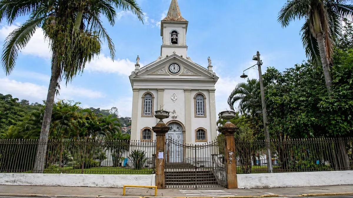 Vista frontal da Igreja Matriz de Santana em Barra do Piraí destacando fachada branca relógio central torre sineira e palmeiras no entorno.