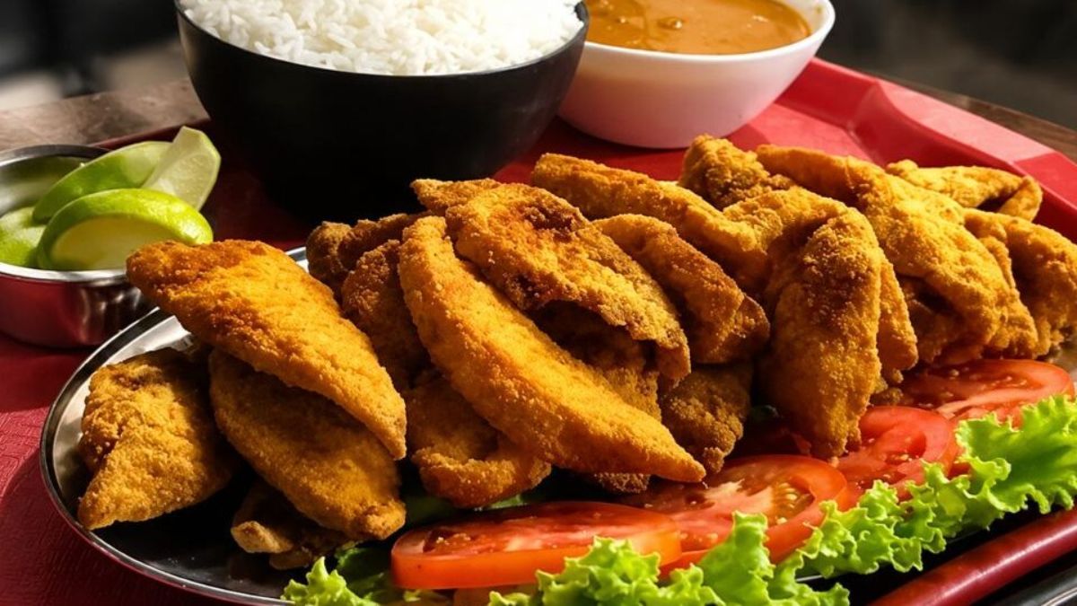 Close em travessa com filés de peixe empanados fritos e dourados servidos com arroz branco pirão de peixe e salada de alface com tomate e limão em cima de uma bandeja vermelha.
