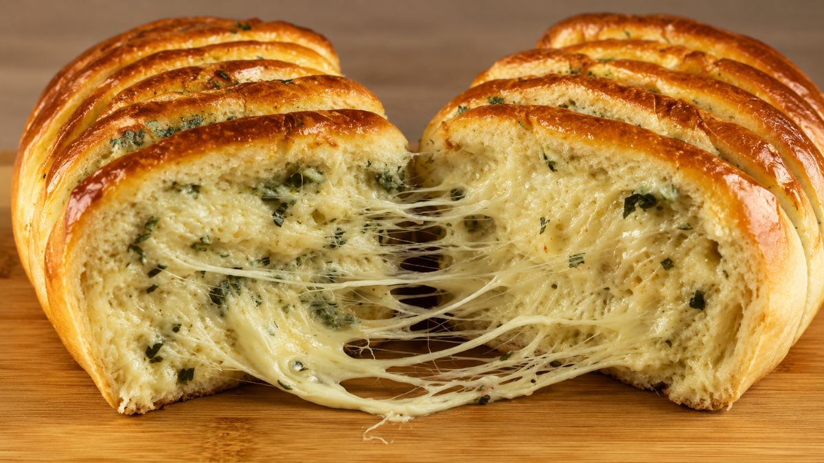 Pão de alho caseiro recheado e fatiado, sendo aberto ao meio com queijo mussarela derretido puxando em fios sobre uma tábua de madeira.