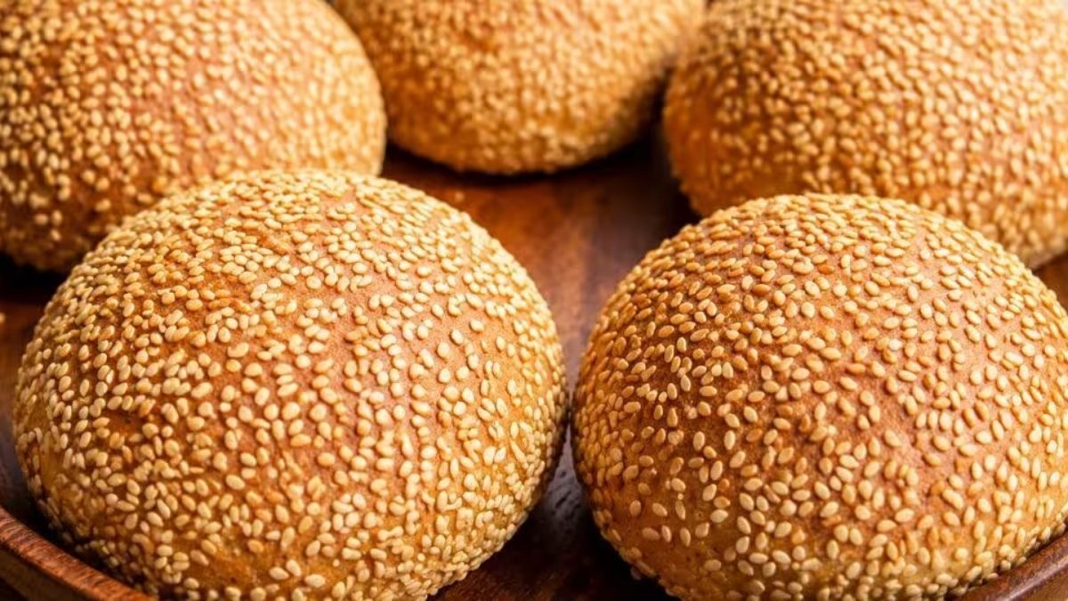 Pães artesanais arredondados cobertos com gergelim torrado dispostos em superfície de madeira, destacando a crosta dourada e a textura fofinha da massa.