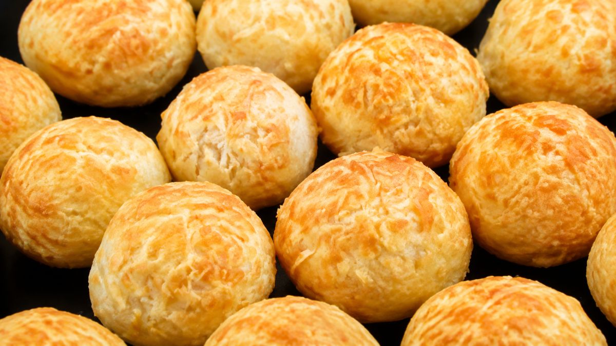 Vários pães de queijo de tapioca redondos e dourados organizados lado a lado sobre uma assadeira escura.