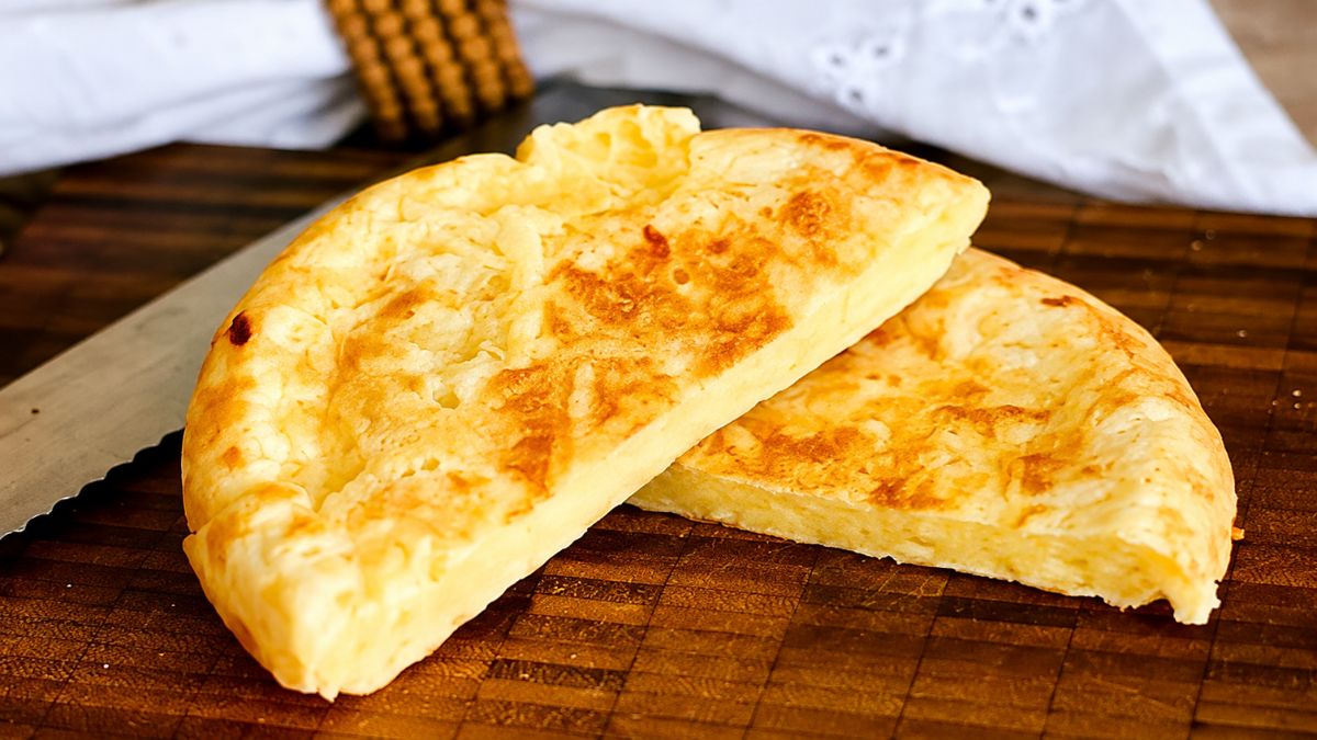Uma foto aproximada foca em duas metades douradas e fofas de um pão de queijo de frigideira caseiro, recheadas com queijo derretido, sobre uma tábua de madeira rústica e com um pano de linho e faca ao fundo.