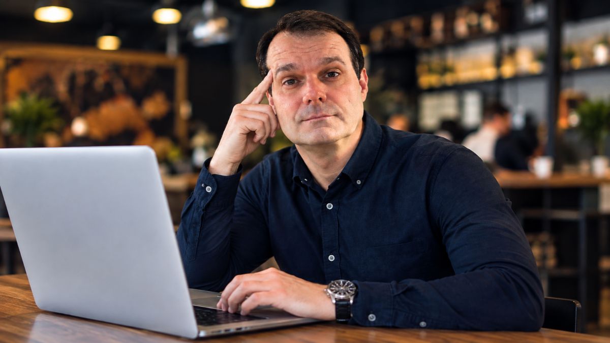 Carlos Jobs sentado à mesa em um ambiente corporativo com notebook aberto, apoiando o indicador na cabeça em atitude pensativa.